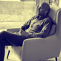 A man sits in a chair with earbuds in listening to something playing on his cellphone.