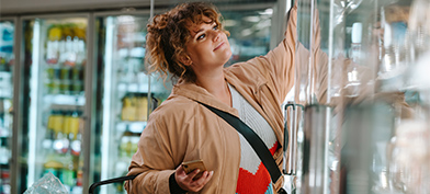 A woman is grocery shopping and reaching into the freezer.