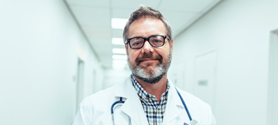 A male doctor smiles at the camera while standing in a hospital hallway.