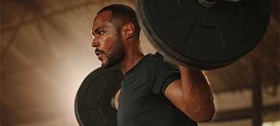 A man holds a bar bell on his shoulders and looks ahead with a concentrated expression. 