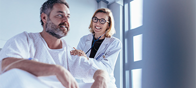 A man sits up in a hospital bed while a doctor listens to his back with a stethoscope. 