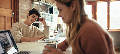 A man looks over at his partner while both are sitting at a table together on their laptops.