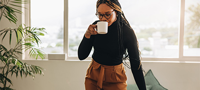 A woman stands at her desk drinking coffee from a mug and looking down at her laptop.