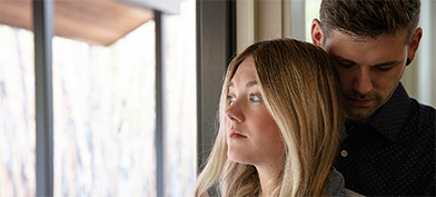 A couple stand by the window with a man hugging a woman from behind.