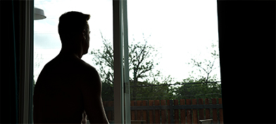 A man living with erectile dysfunction looks out the window in a silhouette. 