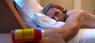 A man lays on his stomach on a bed with his head facing the side and looking sad.