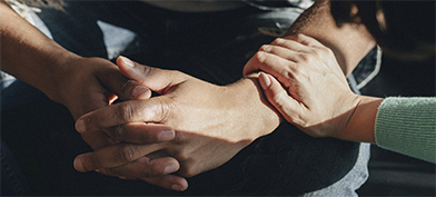 Two hands are folded together with another reaching out to hold his arm on the wrist.