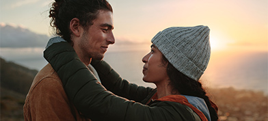 A man and a woman embrace cliffside in front of the sunset.