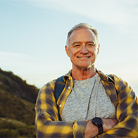 An older man is standing on a mountain and smiling at the camera with his arms folded.