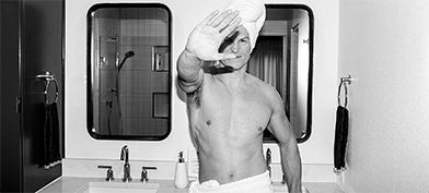 A man in a towel around his waist and on his head holds his hand up to block the camera.