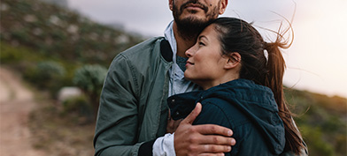 A couple stands outside, the woman leaning her head on a mans chest as he places his hand on her upper arm.