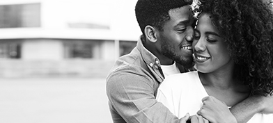 A smiling man wraps his arm around a smiling woman from behind.