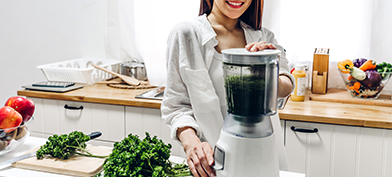 A smiling woman in a kitchen stands next to a cutting board with parsley on it and places her hands on a blender.