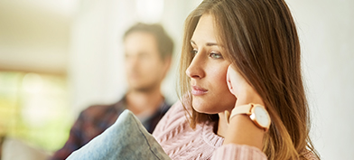 A woman stares blankly ahead with her head resting on her fist and her male partner sitting in the background.