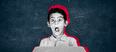 A little boy sits at a desk and makes a fearful expression with his eyes and mouth wide open.