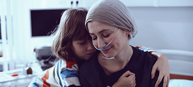 A young child hugs their mother who is wearing a head scarf and oxygen cannula.