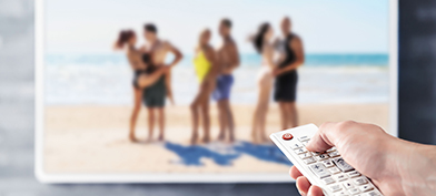 A hand holds a remote pointed at a television with a reality dating show on the screen.