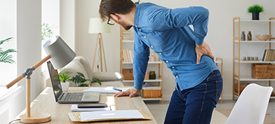 A man holds onto his lower back in pain while leaning on his computer desk.