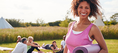 A woman smiles at the camera, wearing a pink tank top and holding an exercise mat.
