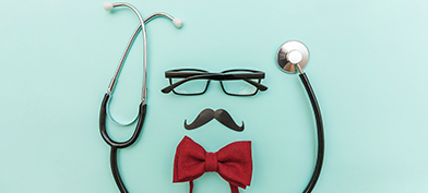 A pair of glasses and a mustache lay against a mint background with a bowtie beneath and surrounded by a stethoscope.
