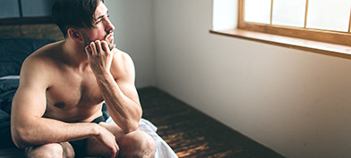 A man sits on the end of his bed scratching his beard and looking out the window.