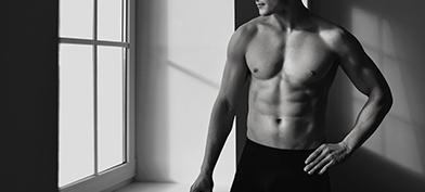 A muscled man in boxer briefs stands shirtless next to a window.