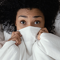 A woman lies in bed with a blanket over the lower part of her face with her eyes wide in fear.