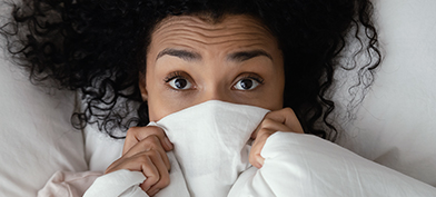 A woman lies in bed with a blanket over the lower part of her face with her eyes wide in fear.