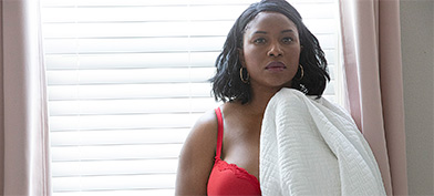 A woman in a red bra holds a white blanket and stands in front of a window with drawn blinds.
