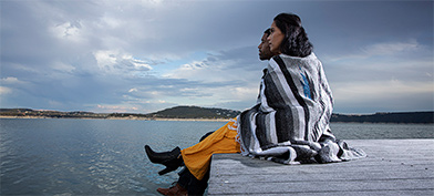 A married couple sits on the edge of a wooden dock looking out over a lake with a blanket around them.