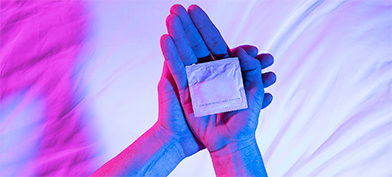 One hand holds the back of another with a wrapped condom in its palm.