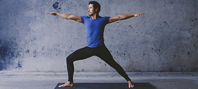 A man in black pants and a blue shirt does the Warrior II yoga pose on a black mat.