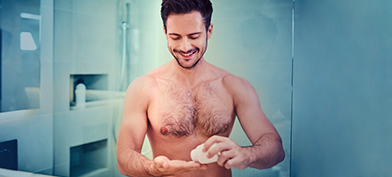 A shirtless man smiles in his bathroom while he pours a pill into his hand from a bottle.