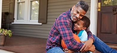A dad sits on a front porch and hugs his child, who is holding a basketball. 