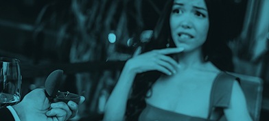 A woman looks nervously at her partner as he holds an engagement ring box in his hand over dinner.