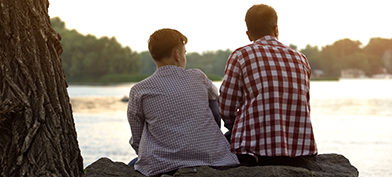 A father and son sitting on a rock face away from the camera and look at a river in front of them.