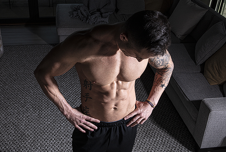 A shirtless man looks downward and holds his hands on his hips.