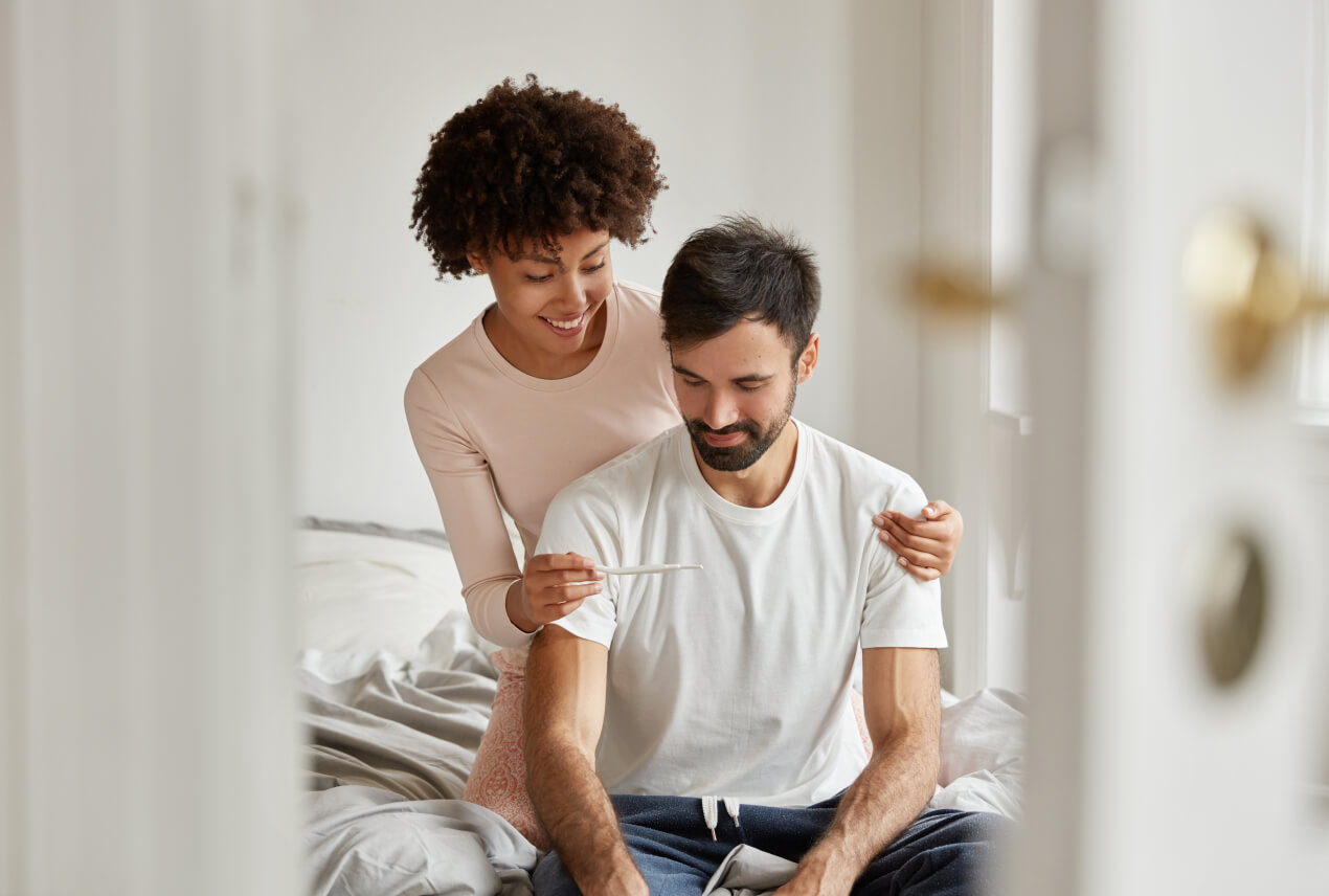 A woman sits behind a man on a bed showing him a pregnancy test and smiling.