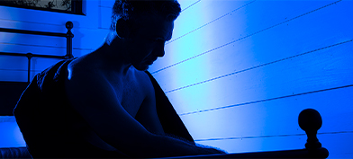 A shadowy figure sits in bed with a blue light shining behind.