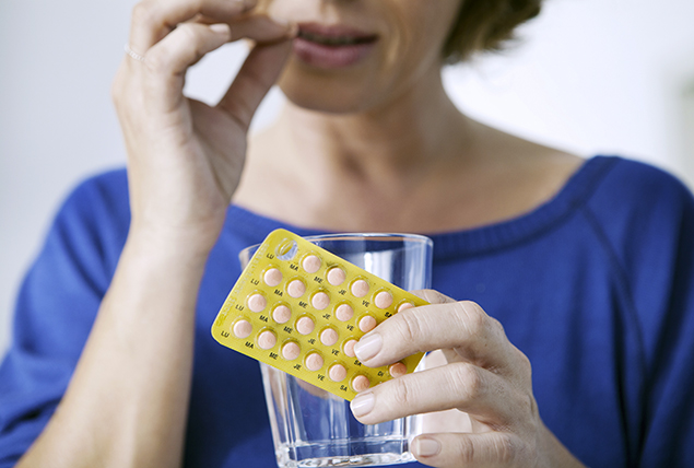 woman holds yellow blister pack of pills and water glass, about to take a pill