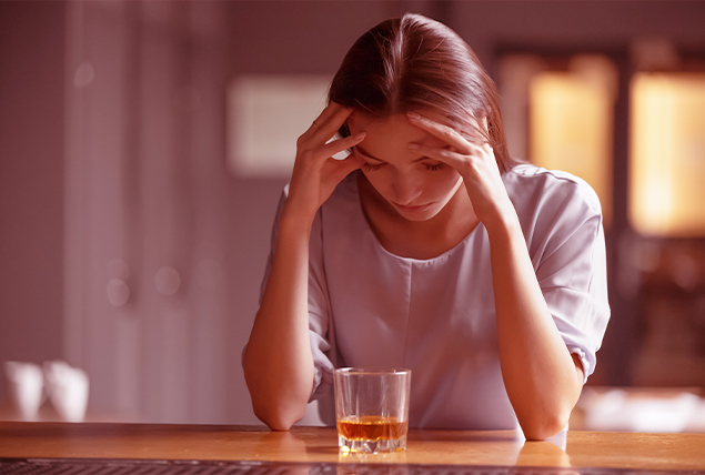A woman holds her head between her hands as she looks down at a glass of whiskey.
