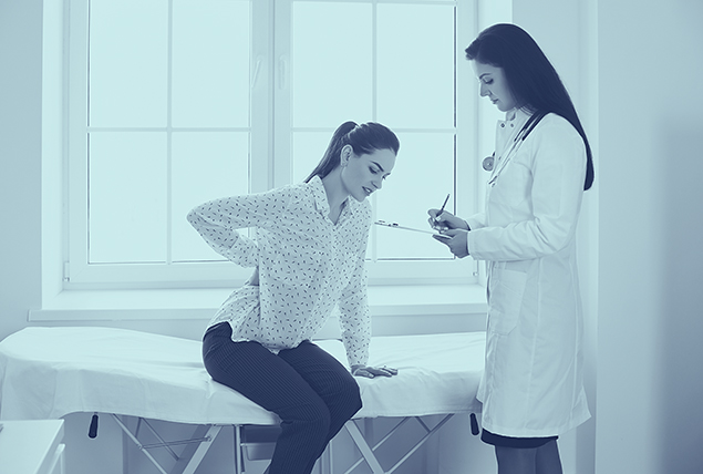 A woman holds her lower back while sitting on a bed as a doctor takes notes on a clipboard. 
