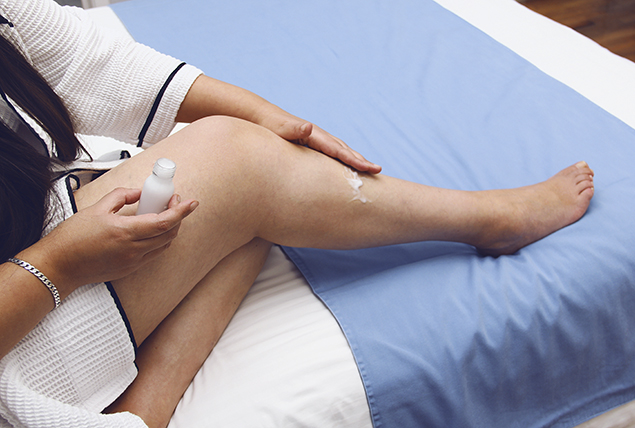 A woman rubs lotion onto her legs while sitting on a bed.