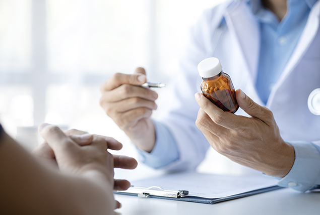 A person folds their hands on a table in front of a doctor holding a pill bottle.