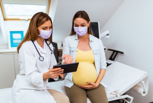 A pregnant woman sits on a hospital bed, looking at a tablet with a doctor.