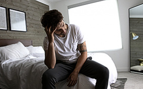 A man sits on the bed holding his head in his hand.