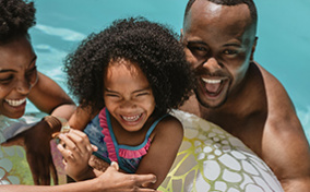 A mother and father laugh as they float in a pool beside their daughter who is in an inner tube.