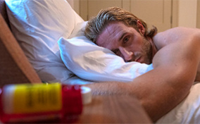 A man lays on his stomach on a bed with his head facing the side and looking sad.