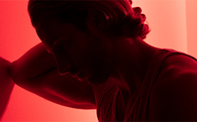 A man leans against a wall while holding his head with a red light shining behind him.