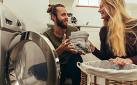 A woman hands her smiling partner laundry from the drying machine.